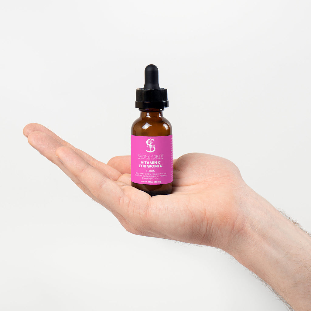 Hand holding a bottle of vitamin C serum with a pink label on a white background, Skinny Pink Fit